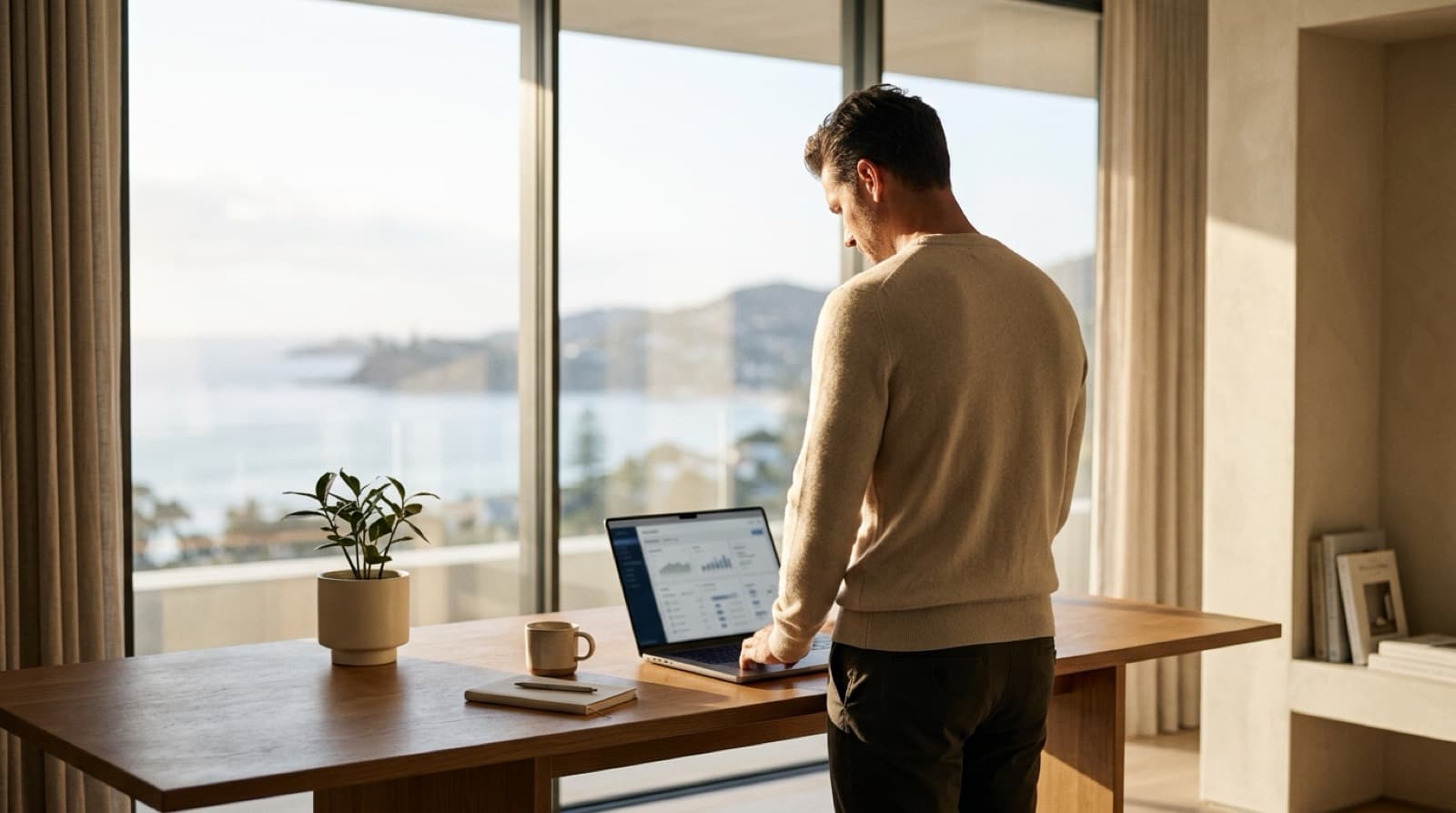 A man standing at a desk, checking a laptop by a window overlooking the sea